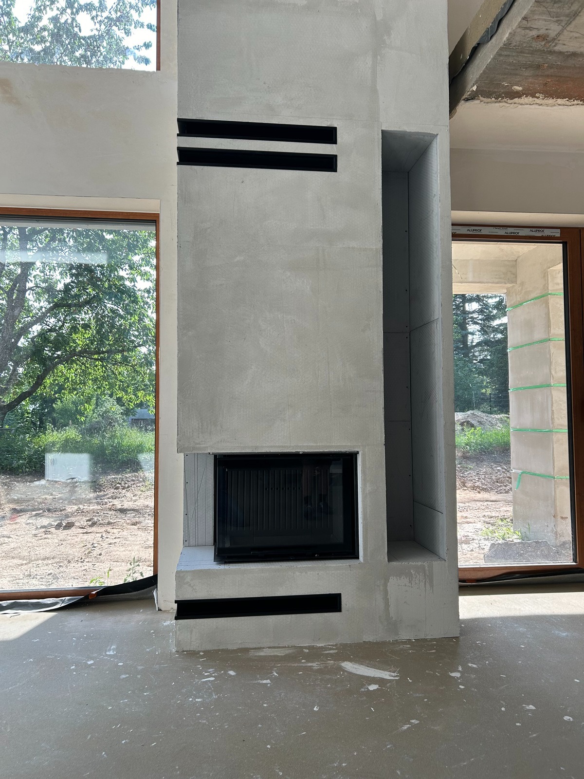 Surowy, betonowy obudowany wkład kominkowy z czarnym frontem i poziomymi szczelinami wentylacyjnymi, w trakcie montażu w nowym budynku z dużymi oknami.