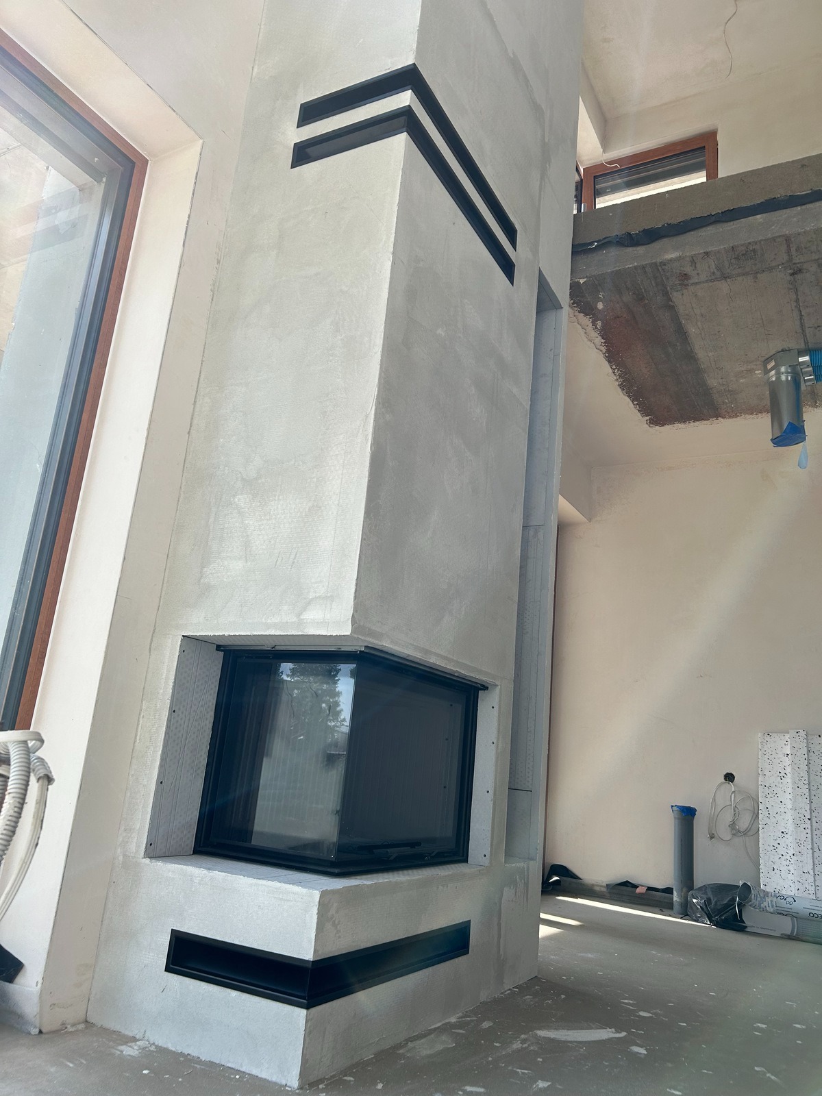 Surowy, betonowy element architektoniczny z czarnymi akcentami, prawdopodobnie obudowa wkładu kominkowego, w trakcie montażu w surowym wnętrzu, widoczne elementy instalacji i okno.