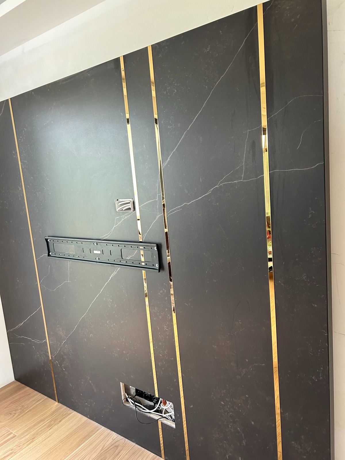 Ściana z ciemnego marmuru z złotymi paskami, uchwyt na telewizor zamontowany, widoczne kable w otworze na dole, przygotowana do montażu kominka elektrycznego.
