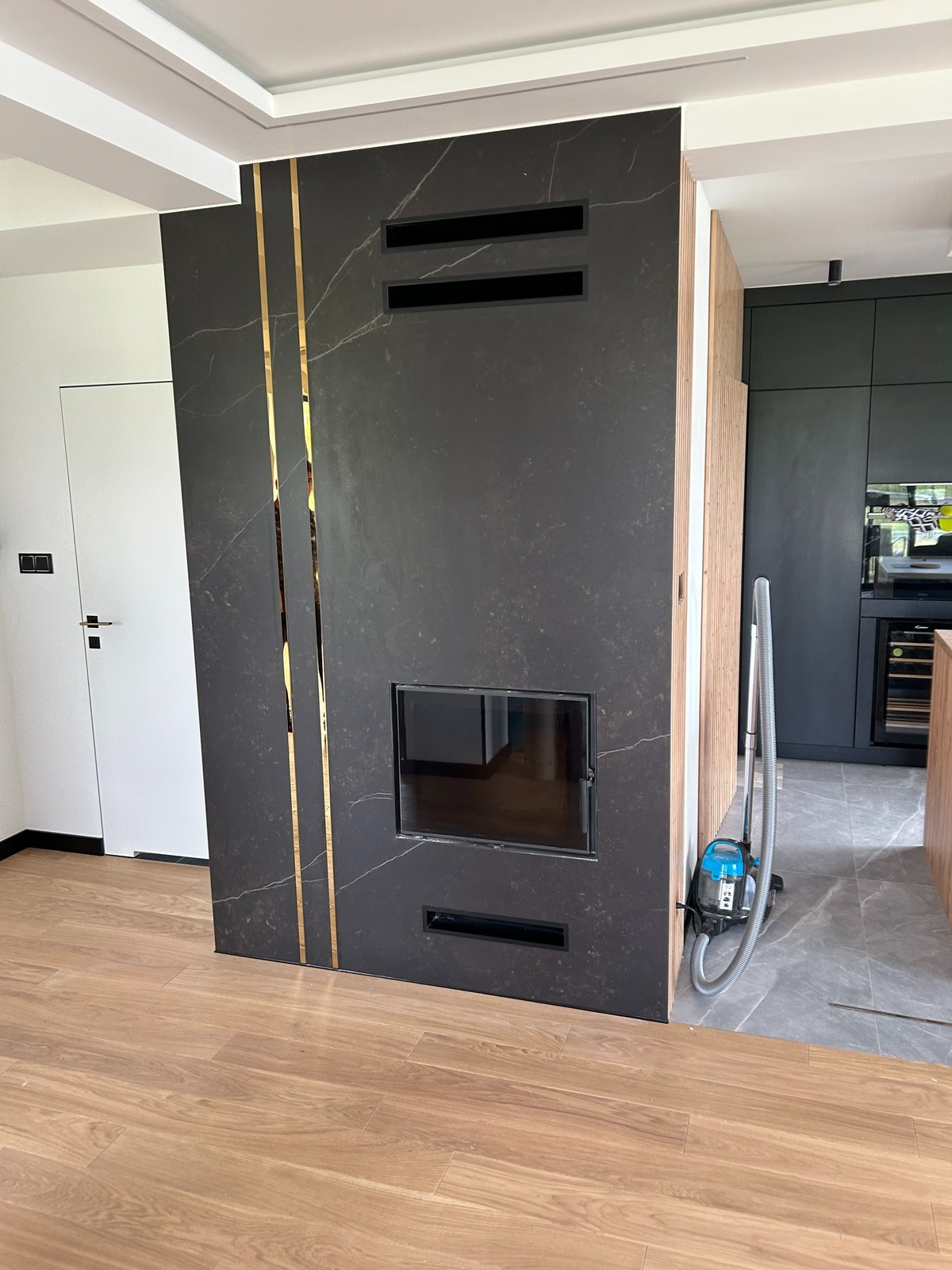 Czarny, marmurkowy portal z wkładem kominkowym, ozdobiony złotymi paskami, z widocznymi kratkami wentylacyjnymi, na tle nowoczesnej kuchni i drewnianej podłogi, z odkurzaczem obok.