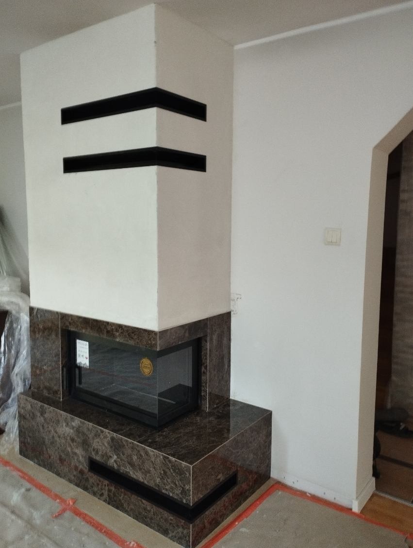 Nowoczesny wkład kominkowy obudowany brązowym marmurem z czarnymi akcentami, widoczne dwa poziome otwory wentylacyjne w białej górnej części obudowy.