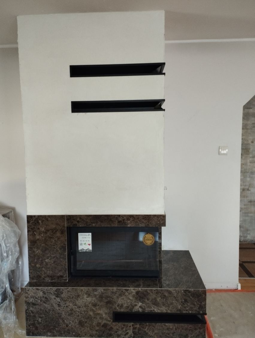 Nowoczesny kominek z obudową z brązowego marmuru, z czarnymi poziomymi wlotami powietrza w górnej części i szybą kominkową z naklejkami informacyjnymi.