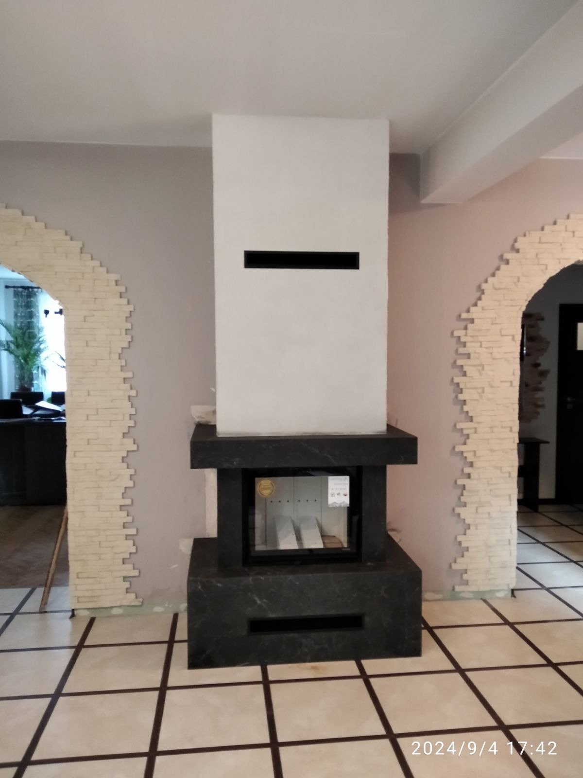 Nowoczesny, minimalistyczny wkład kominkowy z obudową z ciemnego kamienia, wkomponowany w jasne wnętrze z dekoracyjnymi łukami wykończonymi imitacją cegły.