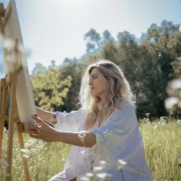 Kobieta w białej koszuli maluje na sztaludze w plenerze, otoczona kwitnącą łąką i drzewami, z promieniami słońca prześwitującymi przez korony drzew.