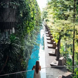 Kobieta w długiej sukience stoi na drewnianym tarasie, opierając się o barierkę i spogląda na długi, wąski basen otoczony zielenią i budynkami porośniętymi roślinnością.