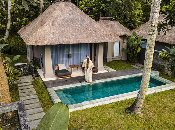 Kobieta w długiej sukience stoi boso przy basenie na tle domku z trzcinowym dachem i leżakiem.