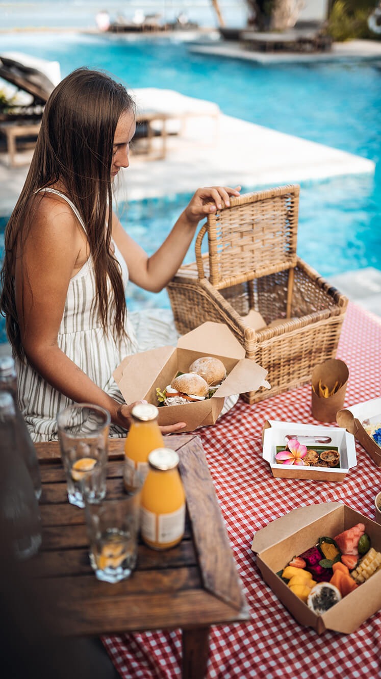 Kobieta otwiera wiklinowy kosz piknikowy z kanapkami, obok napoje i owoce na czerwono-białym obrusie w kratę, w tle basen i leżaki.