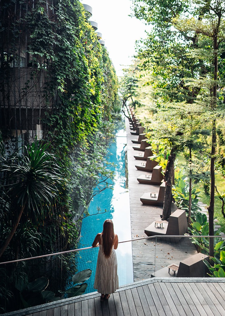 Kobieta w długiej sukience stoi na drewnianym tarasie, opierając się o barierkę i spogląda na długi, wąski basen otoczony zielenią i budynkami porośniętymi roślinnością.