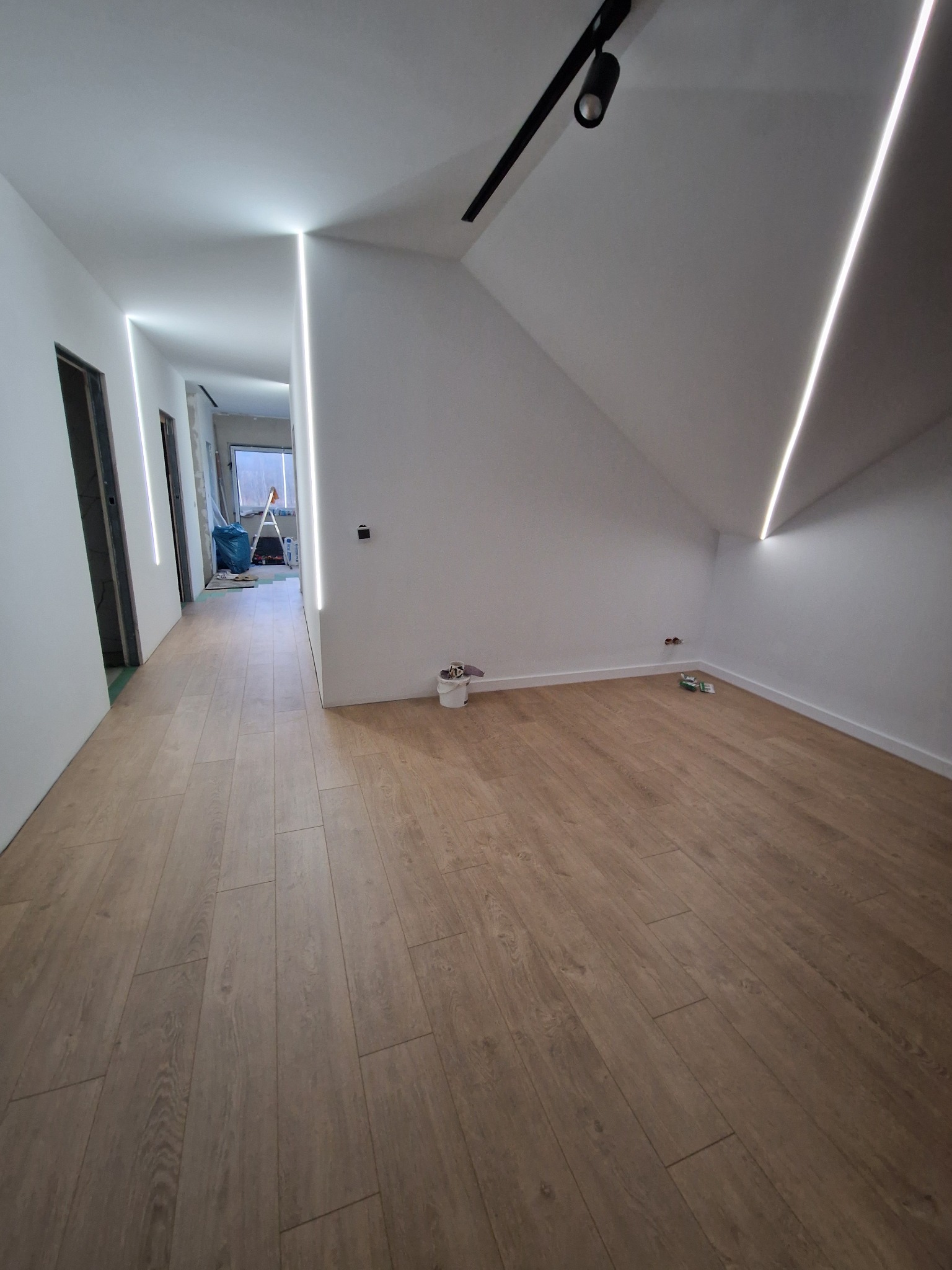 Poddasze po remoncie z drewnianą podłogą, białymi ścianami z liniowym oświetleniem LED i czarnym reflektorem punktowym na suficie; w tle widoczne drabina malarska i niebieski worek.