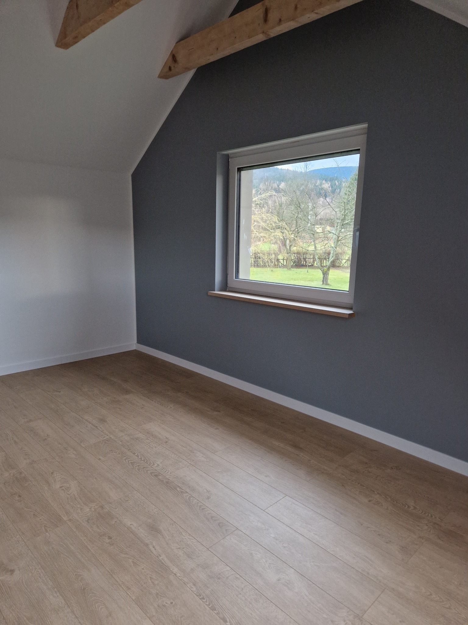 Pokój na poddaszu po remoncie: drewniana podłoga, szara ściana z oknem z widokiem na drzewa, biała ściana, drewniany element konstrukcyjny pod sufitem.