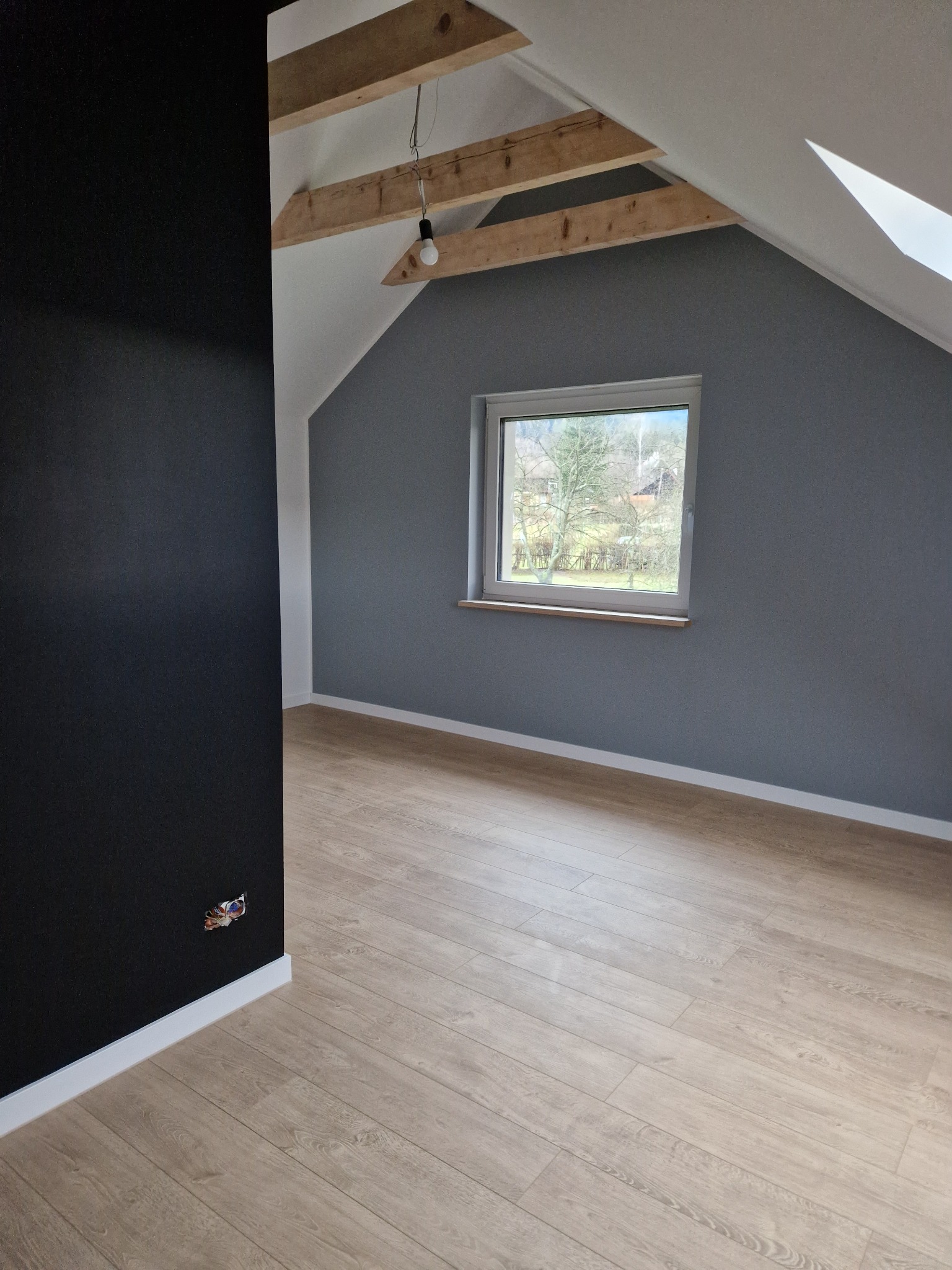 Pokój na poddaszu po malowaniu ścian na szaro i czarno, z drewnianą podłogą, widoczne drewniane belki stropowe i okno z widokiem na drzewa.