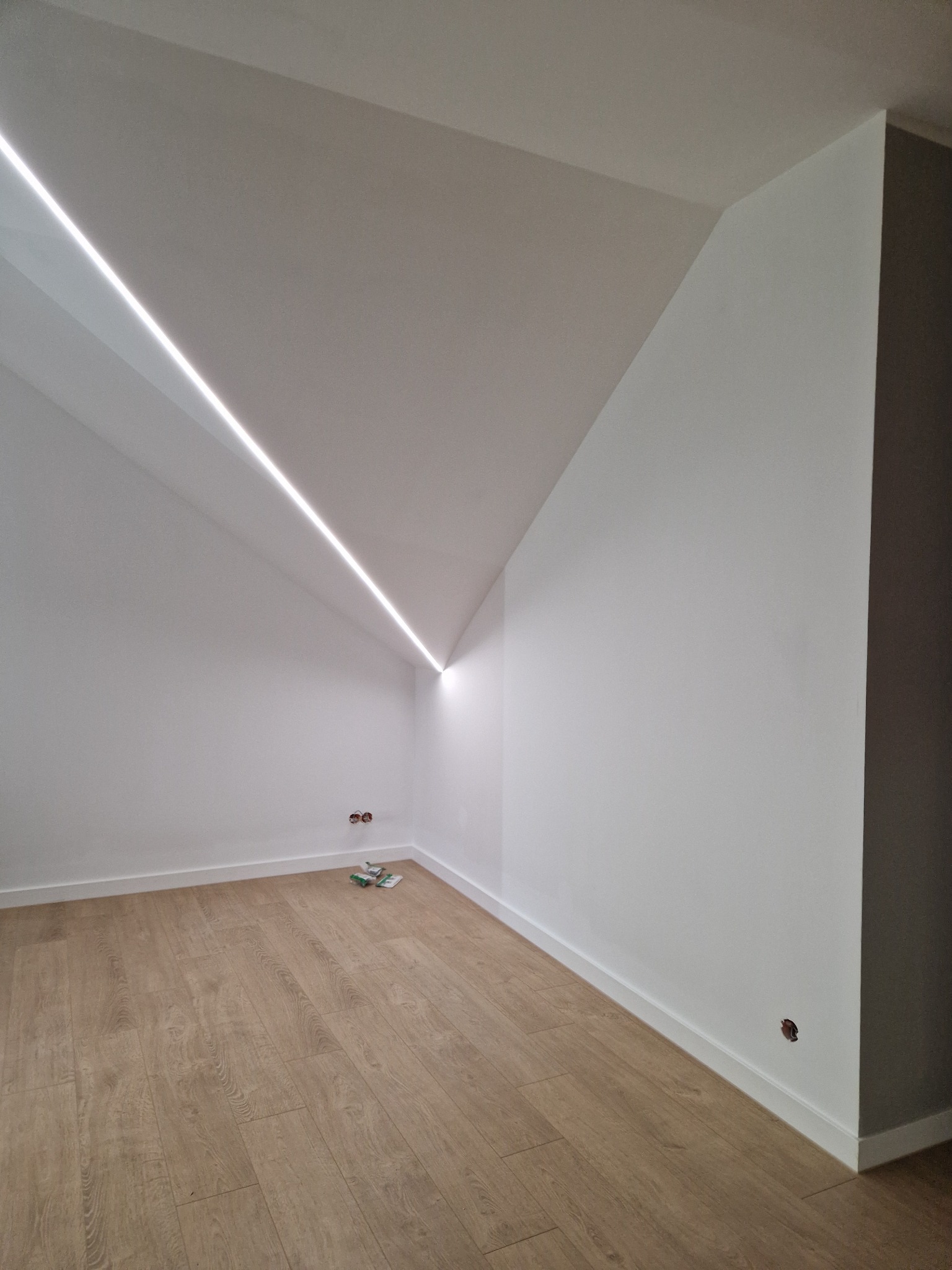 Poddasze po remoncie z oświetleniem LED w suficie, drewniana podłoga, białe ściany i listwy przypodłogowe, widoczne puszki elektryczne i resztki materiałów budowlanych.