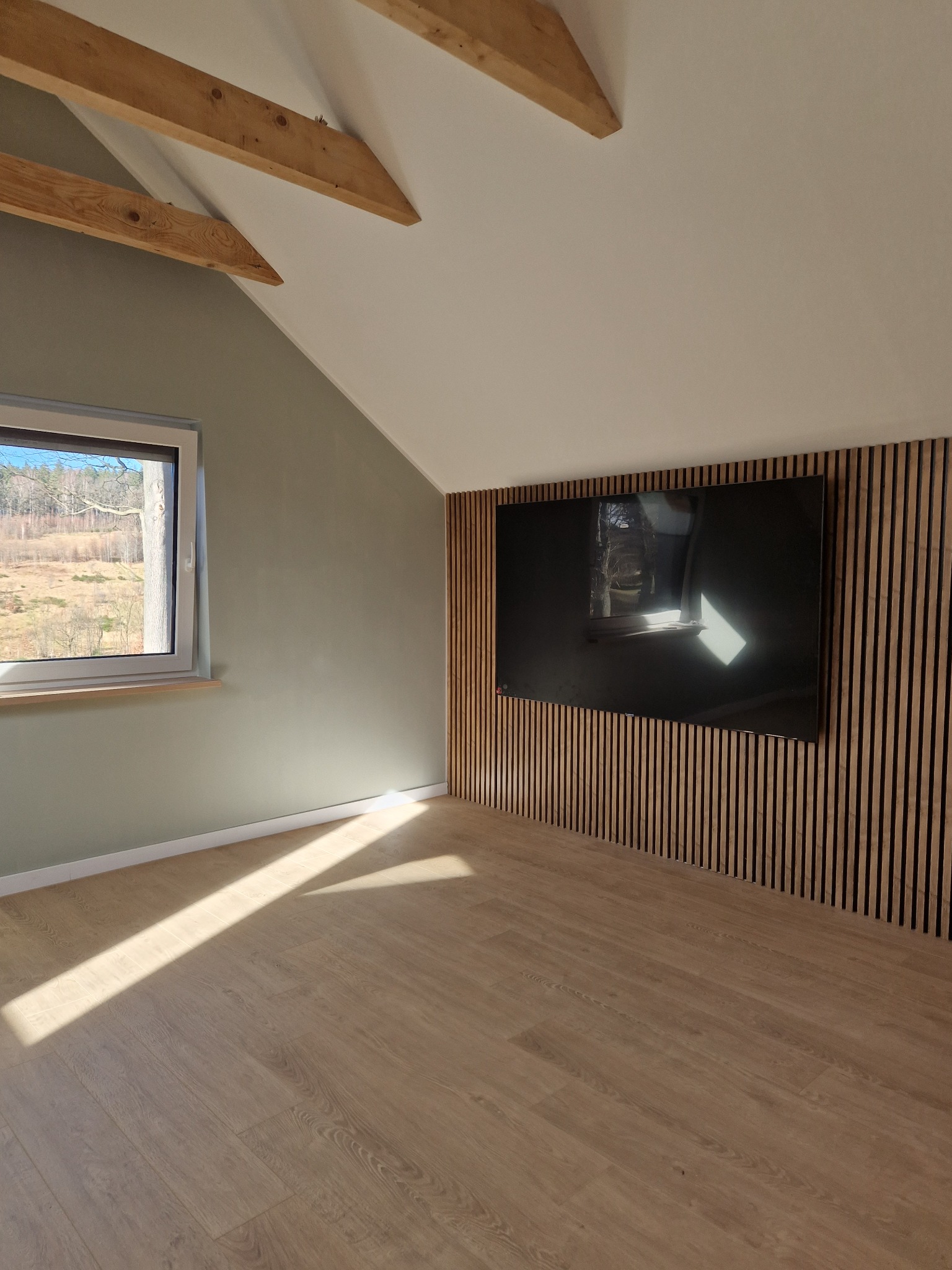 Wykończone poddasze ze skośnym sufitem, drewnianą boazerią za telewizorem i parkietem, oświetlone naturalnym światłem wpadającym przez okno.