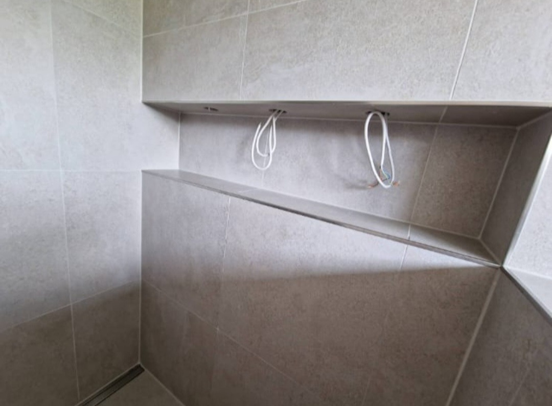 Ściana łazienki wyłożona beżowymi płytkami, z półką wnękową, z której wystają przewody elektryczne