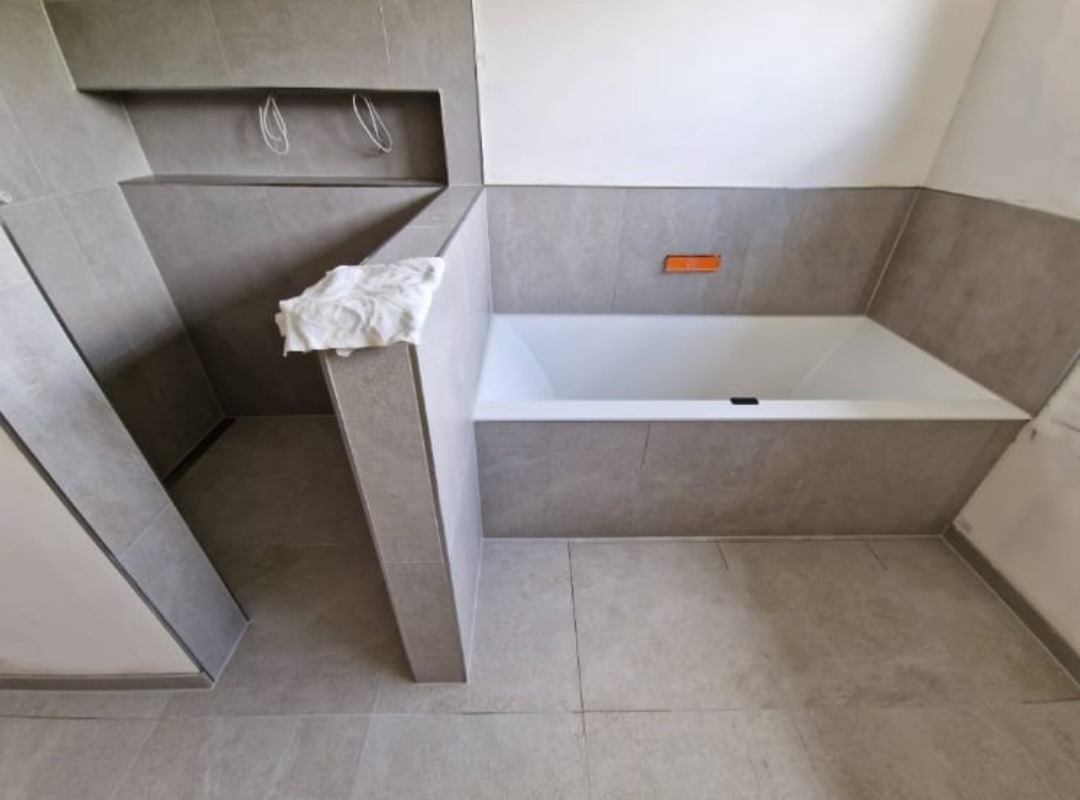 Wanna w obudowie z szarych płytek, z wnęką i ręcznikiem na ścianie, widoczne kable w ścianie, szara podłoga z płytek.