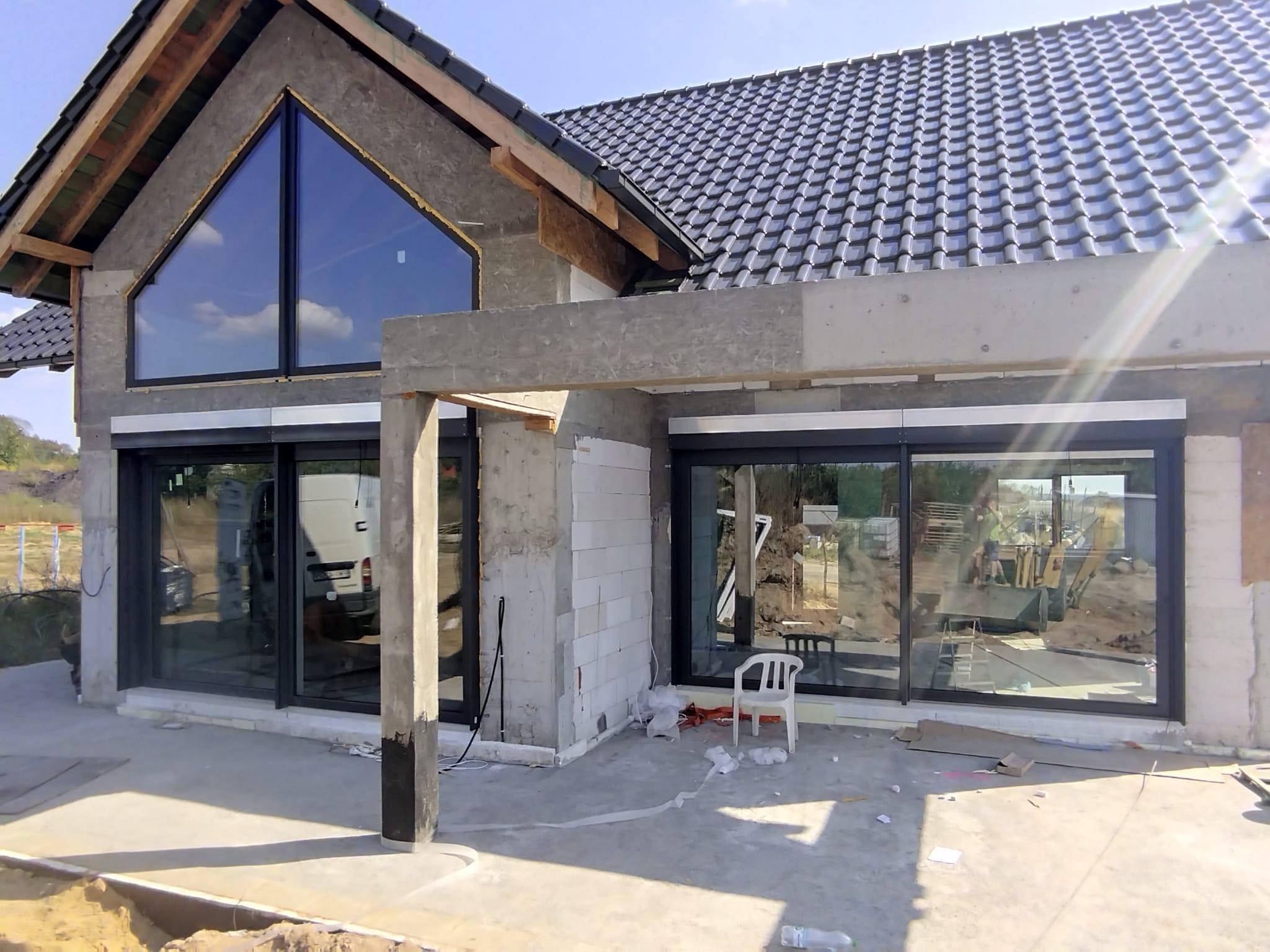 Dom w trakcie budowy z zamontowanymi dużymi przesuwnymi oknami z czarnymi ramami i trójkątnym oknem na poddaszu, widoczny biały bus i koparka w odbiciu szyb.