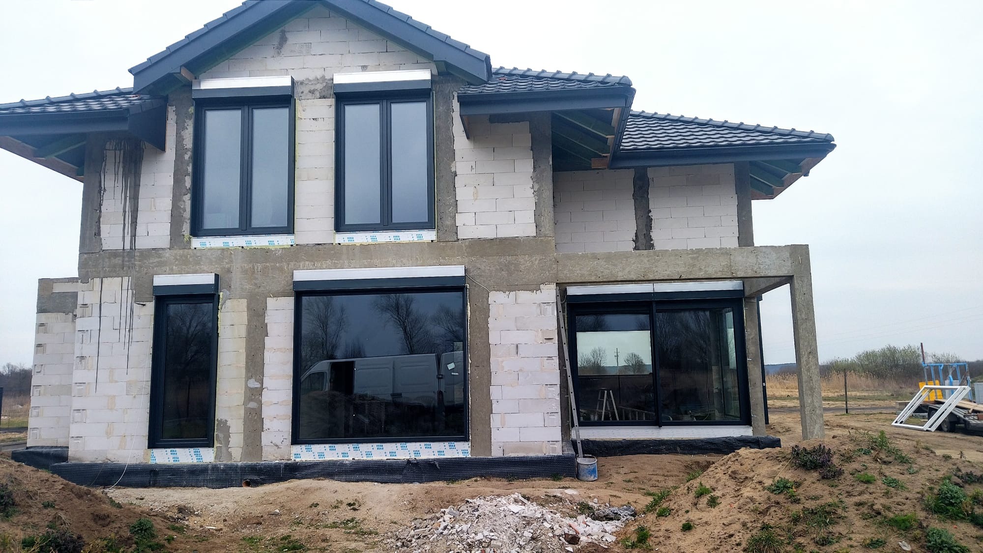 Surowy stan domu w trakcie budowy z zamontowanymi czarnymi oknami, widoczne białe bloczki, ocieplenie i drabina oparta o ścianę.