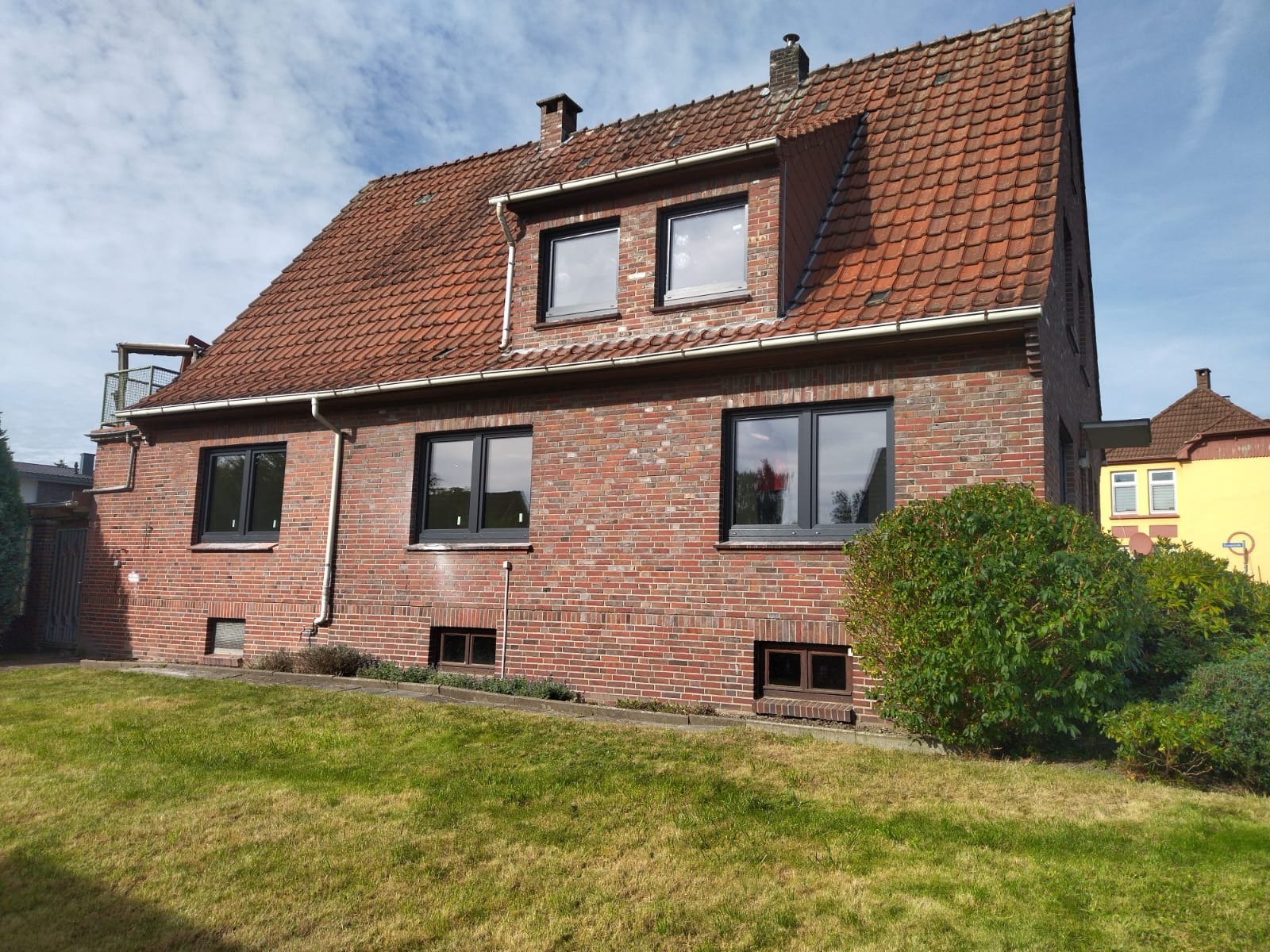 Ceglany dom jednorodzinny z nowymi oknami PCV w czarnych ramach, widoczne z zewnątrz, z trawnikiem z przodu i fragmentem sąsiedniego budynku w tle.