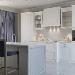 Jasna, klasyczna kuchnia z wyspą, wyposażona w białe szafki z frezowanymi frontami, szklane witryny, okap i wbudowany piekarnik. Na wyspie kuchennej znajdują się dwa ciemnoszare hokery. Za oknem...