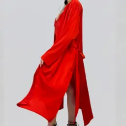 Modelka w czerwonej, satynowej sukience z głębokim dekoltem i rozcięciem, prezentuje zwiewny krój na jasnym tle. Widoczny fragment czarnego naszyjnika.