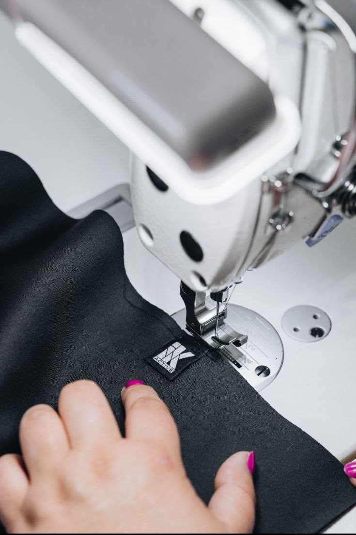 Czarna tkanina z przyszytą metką 'IK ACTIVE' pod igłą maszyny do szycia, dłoń z pomalowanymi na różowo paznokciami stabilizuje materiał.