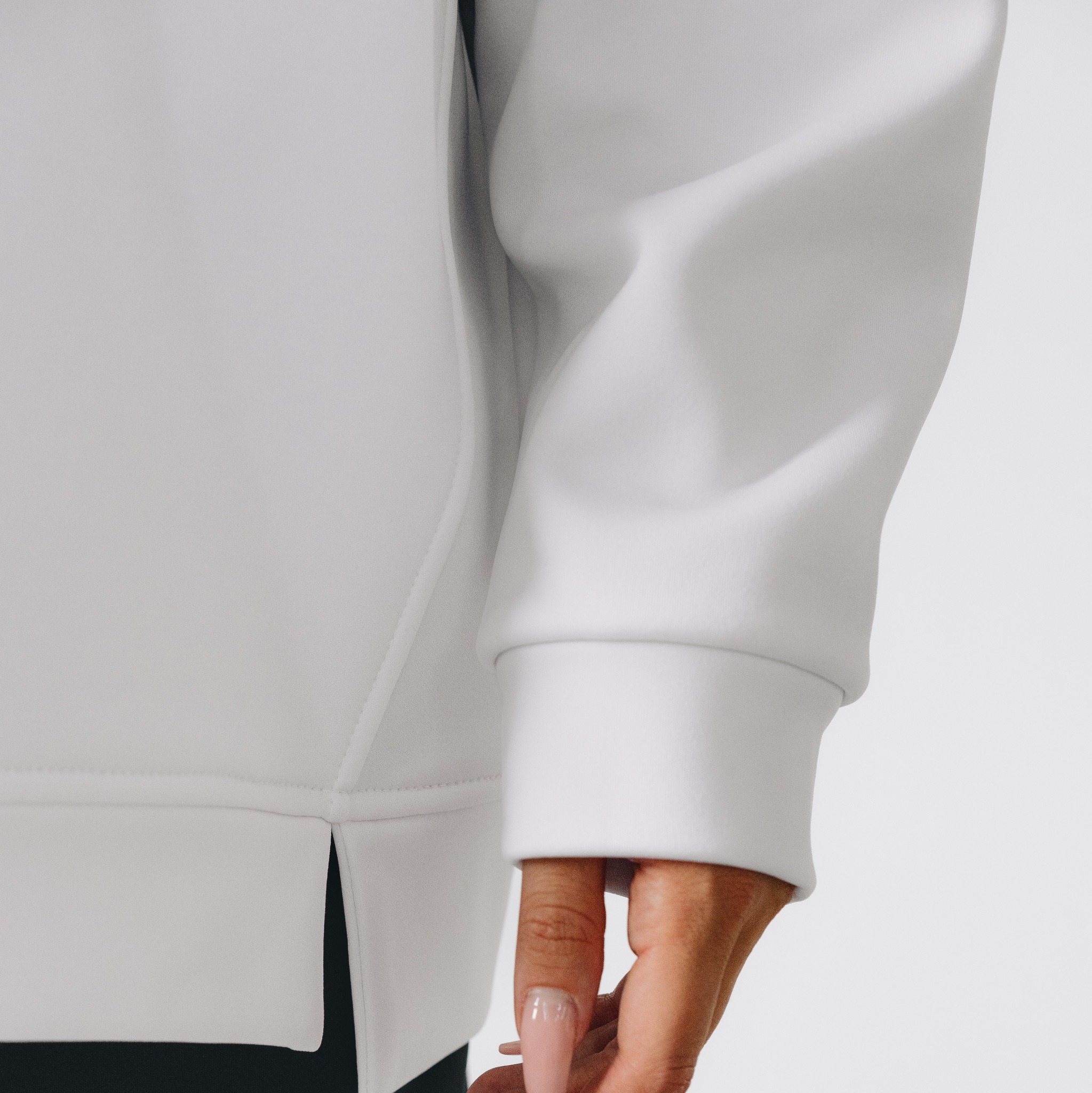 Fragment białej bluzy dresowej z długim rękawem, widoczny detal mankietu i rozcięcia u dołu, na dłoni widoczne długie paznokcie w jasnym odcieniu różu.