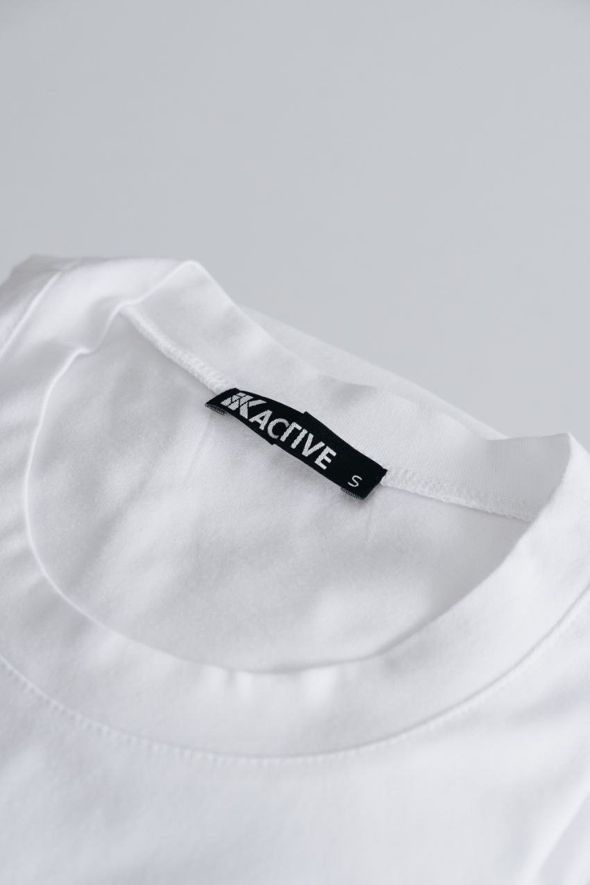 Close-up of a plain white t-shirt with a black label reading 'XACTIVE' and the size 'S', showcasing the neckline and fabric texture.