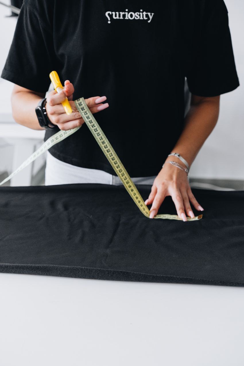 Osoba w czarnej koszulce Curiosity mierzy czarny materiał żółtą miarką krawiecką na białym stole.