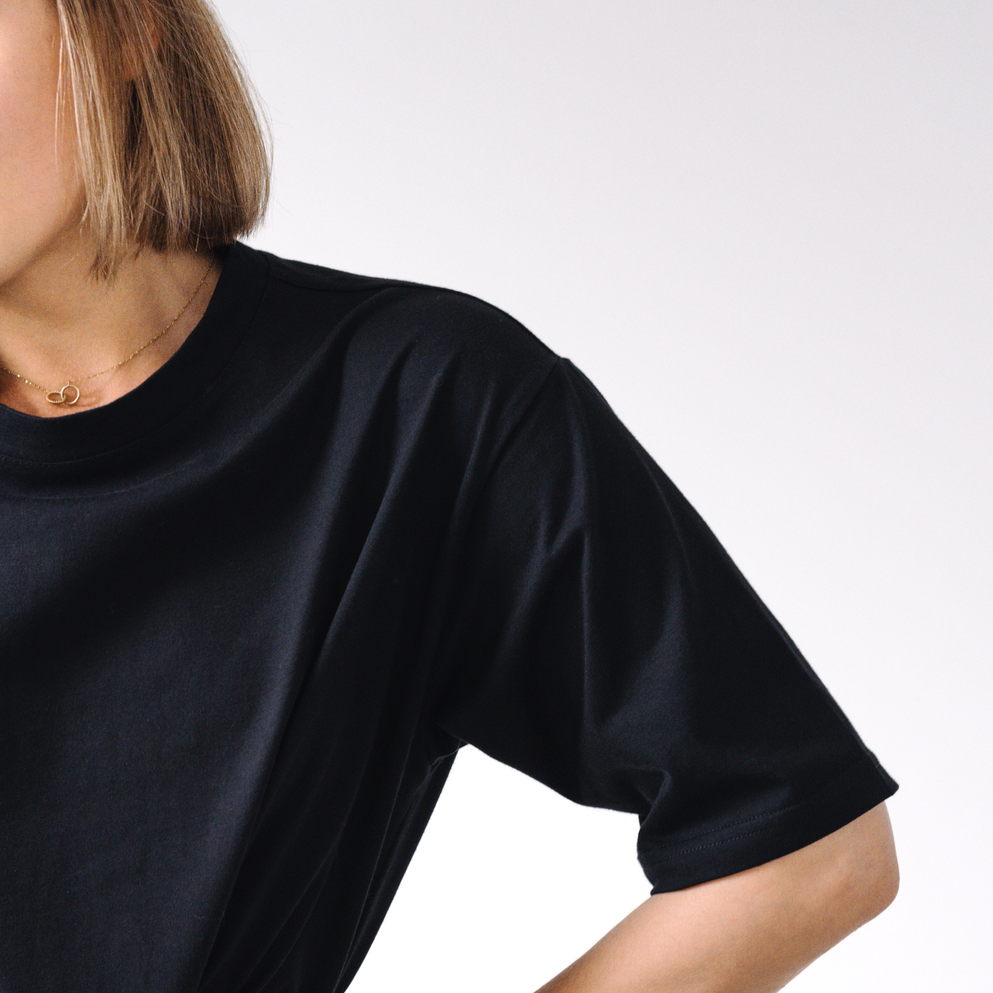 Fragment sylwetki kobiety w czarnym, gładkim T-shircie z krótkim rękawem, widoczny złoty naszyjnik z drobnym wisiorkiem na tle jasnego, jednolitego tła.