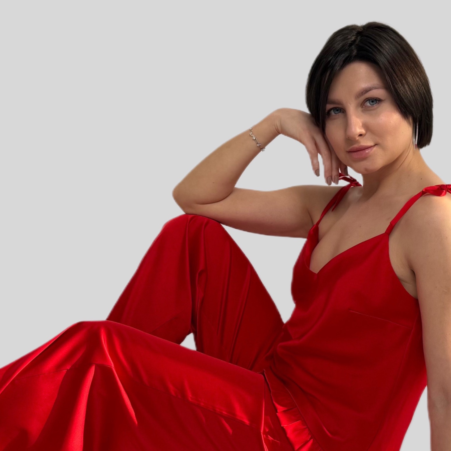 Kobieta w czerwonej satynowej piżamie z topem na ramiączkach i spodniami, pozująca na szarym tle, z bransoletką na nadgarstku i krótkimi, ciemnymi włosami.