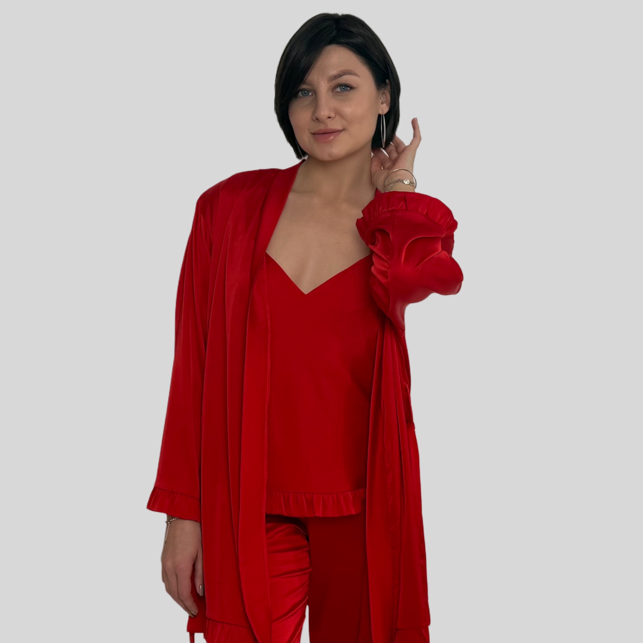 Kobieta prezentuje czerwony satynowy zestaw do spania: top na ramiączkach, długie spodnie i szlafrok. Uśmiechnięta modelka dotyka włosów, na nadgarstku widoczna bransoletka.