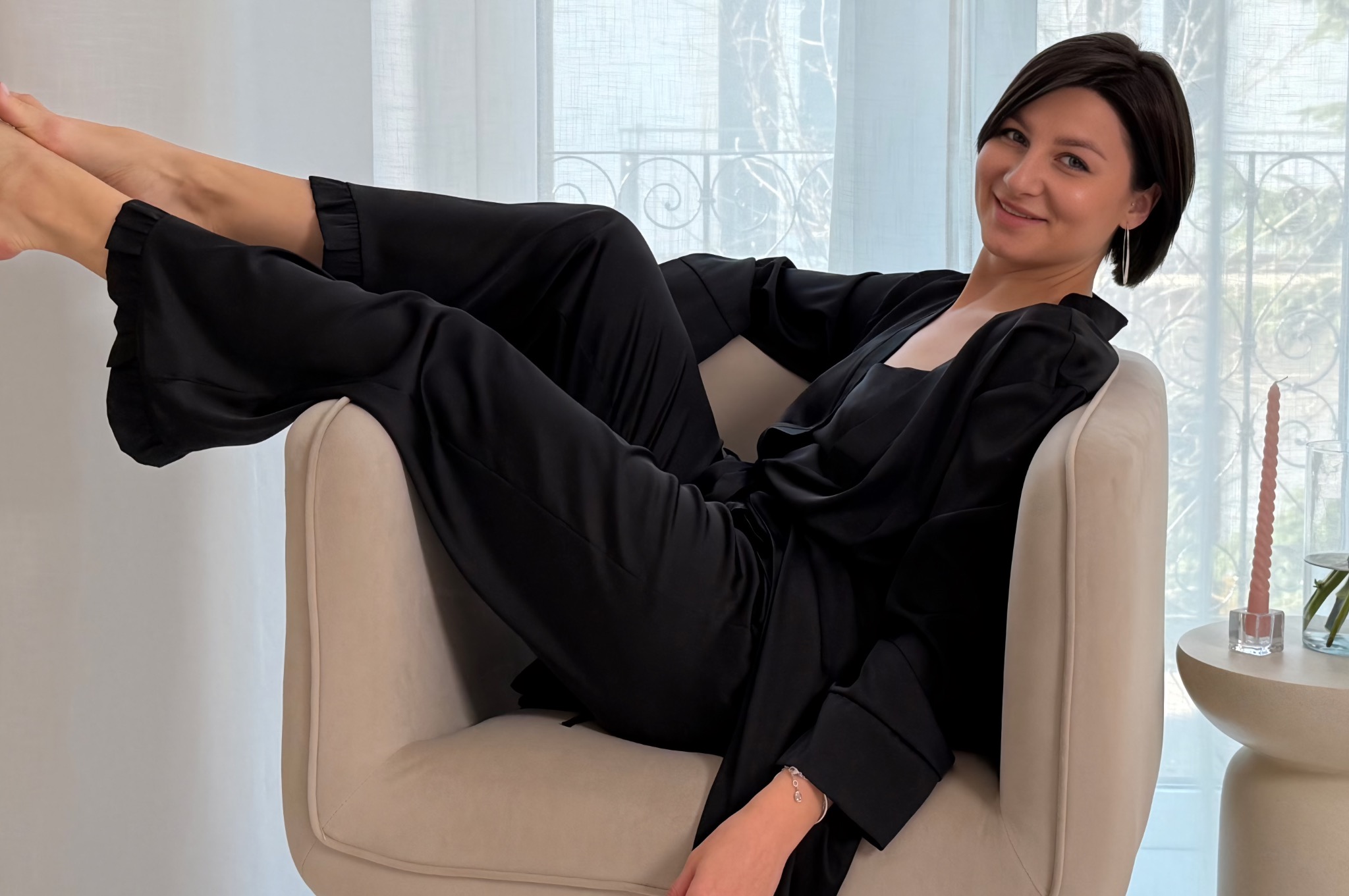 Uśmiechnięta kobieta w czarnym, satynowym komplecie relaksuje się w beżowym fotelu, z nogami uniesionymi do góry, w tle okno z delikatnymi firankami i mały stolik z dekoracją
