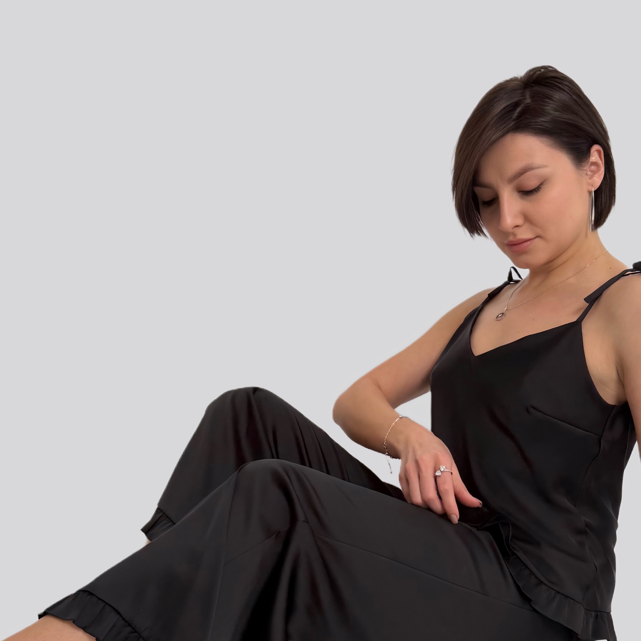 Kobieta siedzi w czarnej satynowej piżamie z falbankami, z delikatną biżuterią, na jasnoszarym tle.