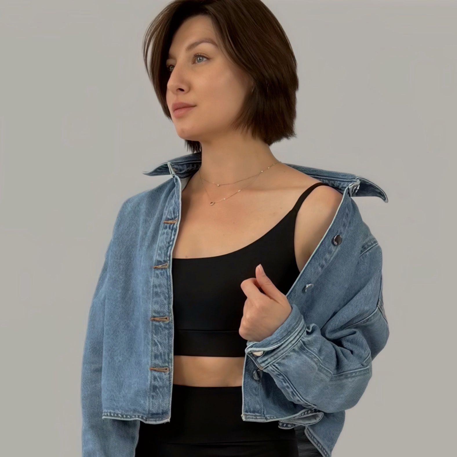 Kobieta pozuje w czarnym topie i jeansowej kurtce, prezentując stylizację. Widoczna biżuteria na szyi i krótka fryzura, neutralne tło.