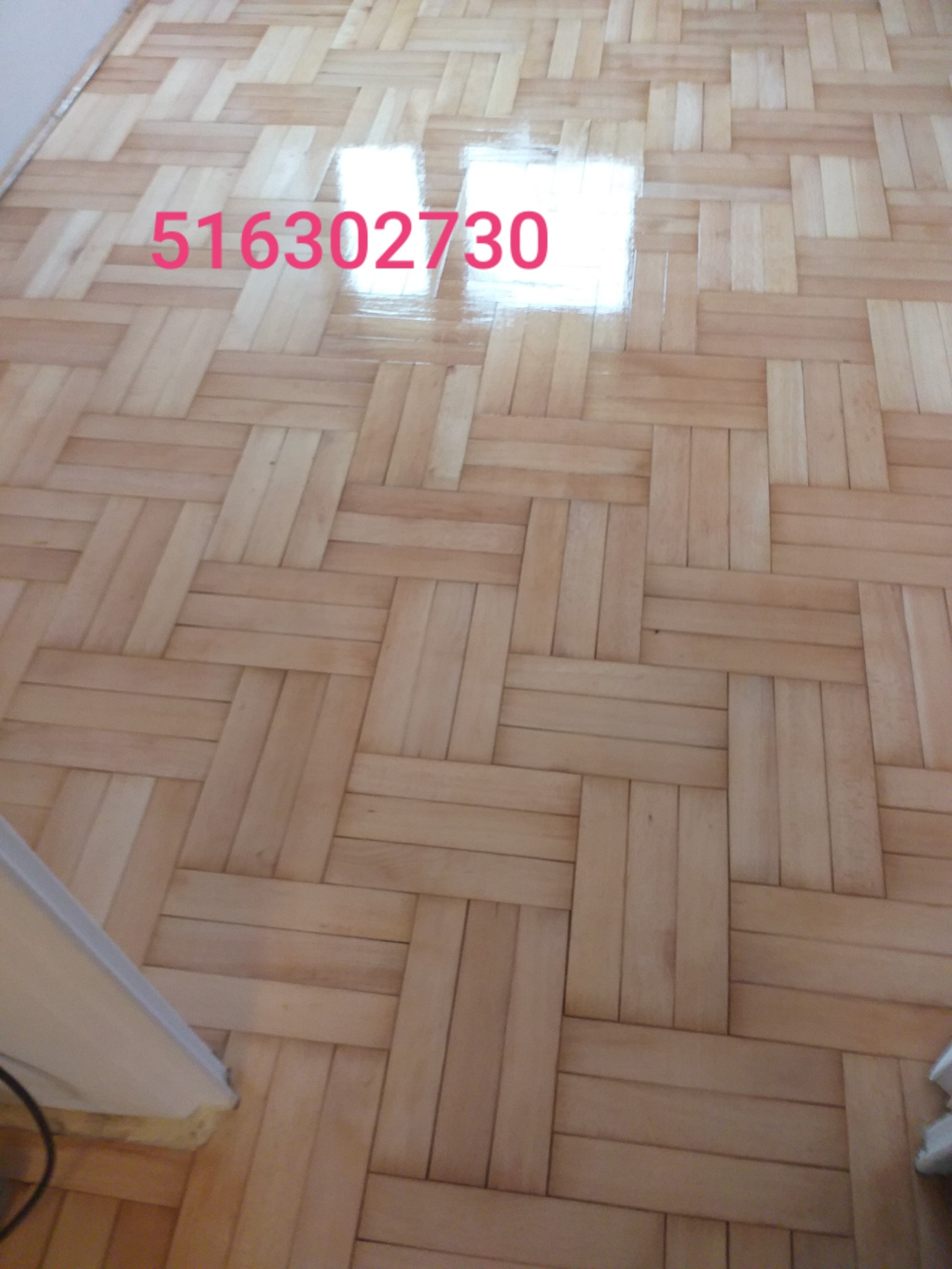 Ułożony parkiet z jasnego drewna w wzór jodełki, z widocznym połyskiem świeżo nałożonego lakieru, fragment ściany w lewym dolnym rogu.