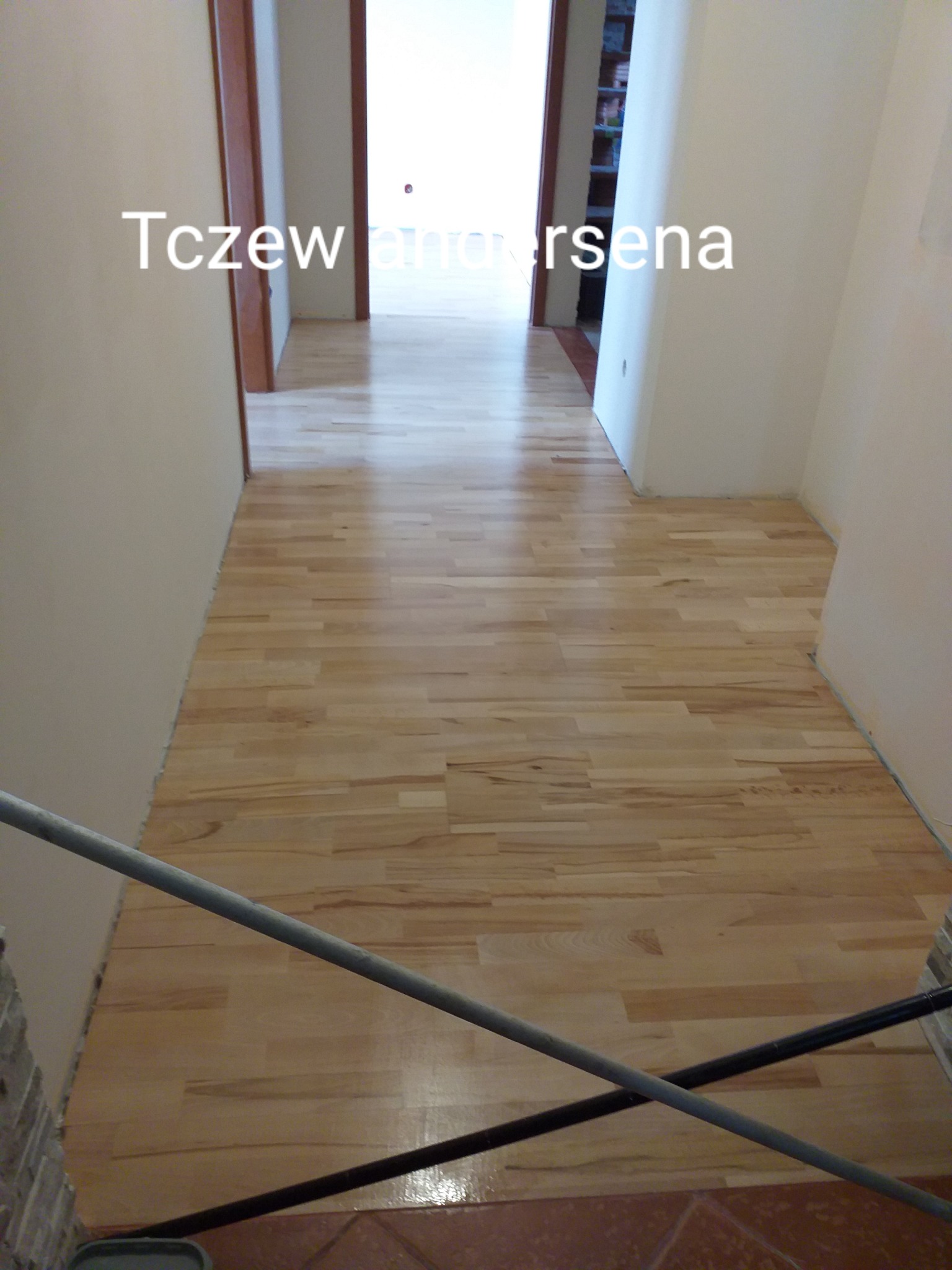 Jasna, świeżo ułożona podłoga z parkietu w korytarzu, widoczne niedoskonałości przy ścianach wymagające wykończenia, w tle otwarte drzwi do kolejnych pomieszczeń.