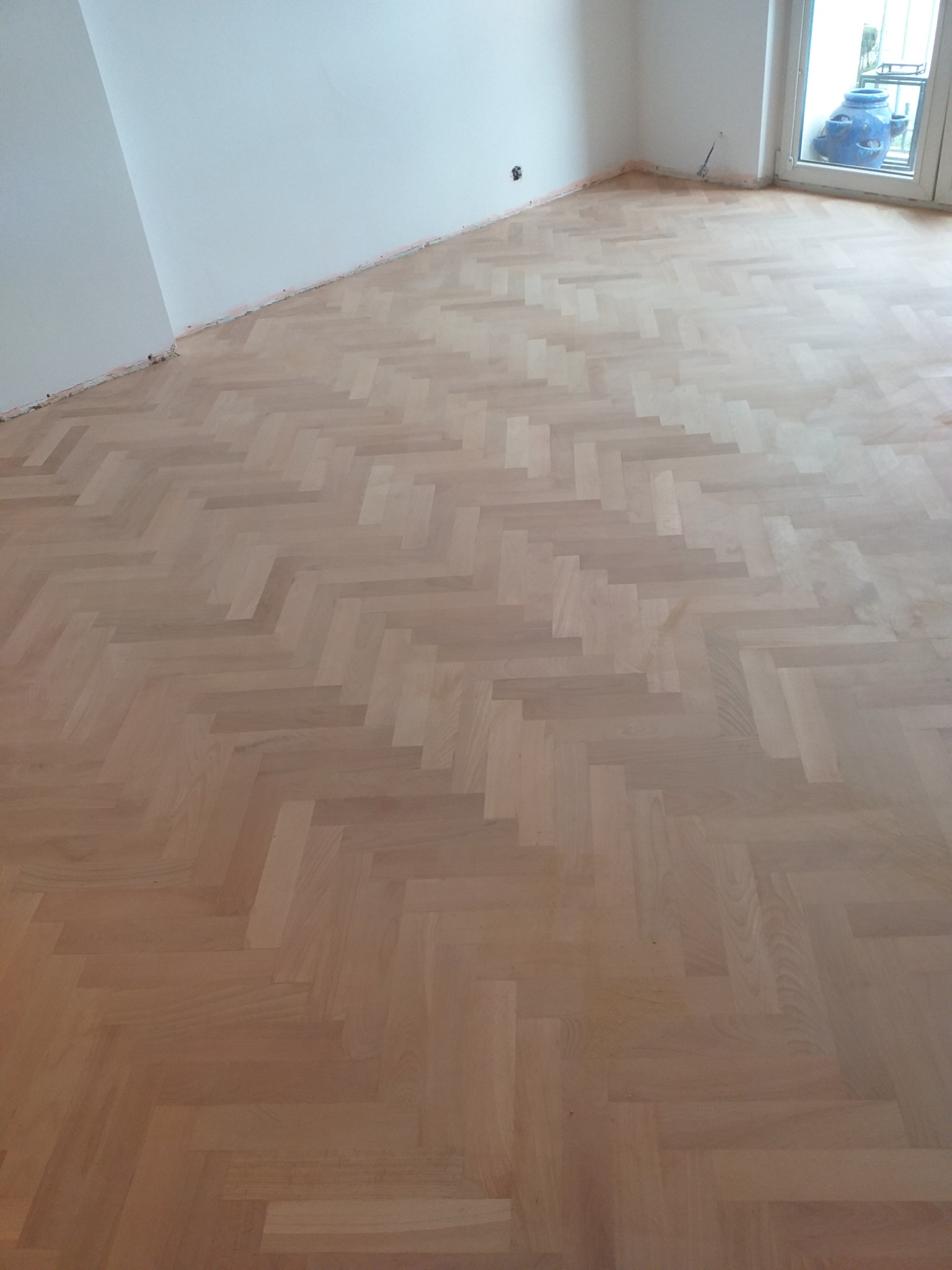 Jasny parkiet ułożony we wzór jodełki w pustym pokoju z białymi ścianami, widoczne ślady po listwach przypodłogowych, w tle okno z dużym wazonem.