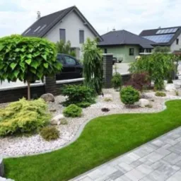 Starannie zaprojektowany ogród przydomowy z równo przystrzyżonym trawnikiem, ozdobnymi krzewami i drzewami o kulistych koronach, wysypany białym żwirem, otoczony szarą kostką brukową i ceglanym murem.