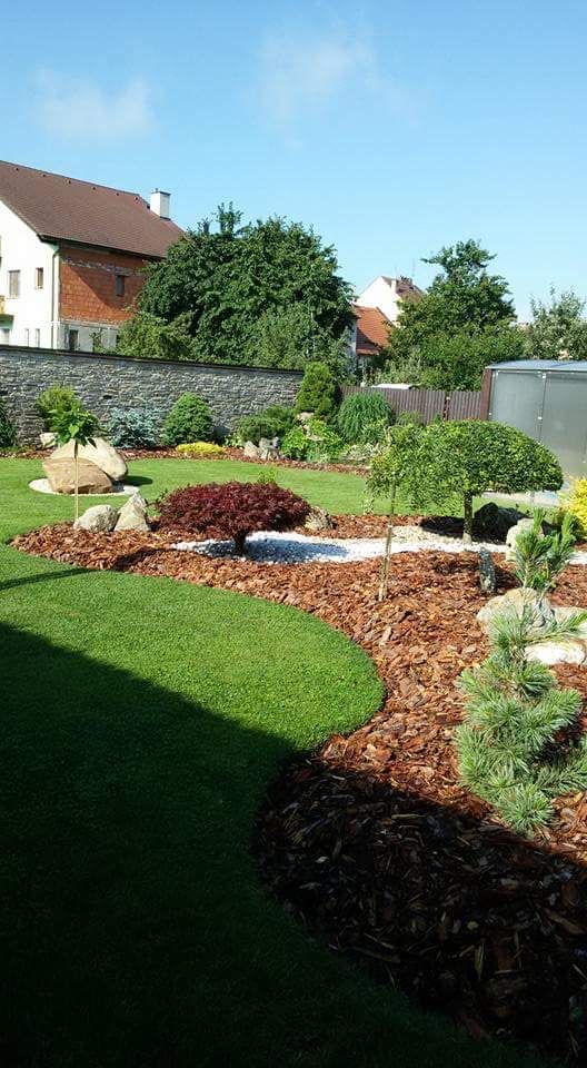 Starannie zaprojektowany ogród z równo przystrzyżonym trawnikiem, ozdobnymi rabatami wysypanymi korą, kamieniami i niskimi drzewami o różnym pokroju i kolorze liści, na tle kamiennego muru...