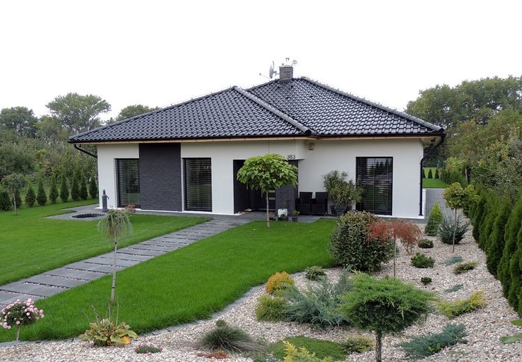 Nowoczesny dom jednorodzinny z zadbanym ogrodem: trawnik, ścieżka z płyt, rabata z ozdobnymi krzewami i drzewami.