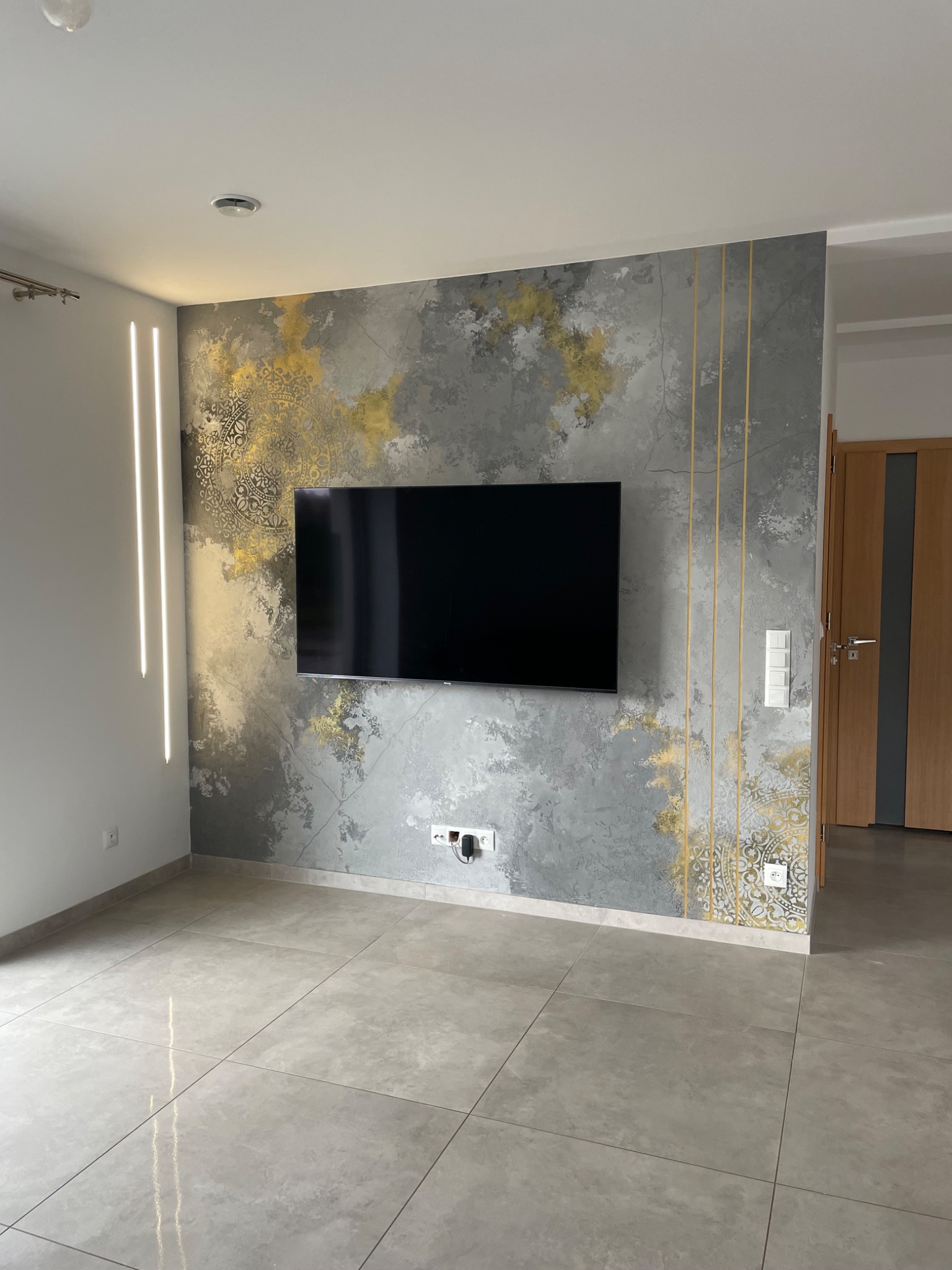 Ściana w salonie z dekoracyjnym tynkiem w odcieniach szarości i złota, z pionowymi złotymi paskami, podwieszony telewizor, podświetlenie LED po lewej stronie, płytki podłogowe w odcieniach szarości.