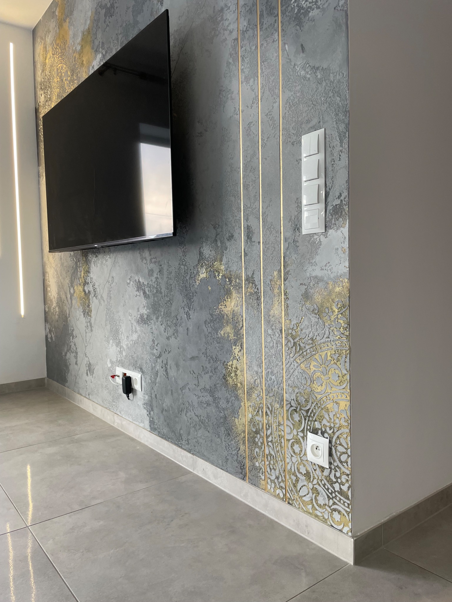 Dekoracyjna ściana w salonie z efektem betonu, złotymi paskami i ornamentem, z zamontowanym telewizorem i podświetleniem LED.