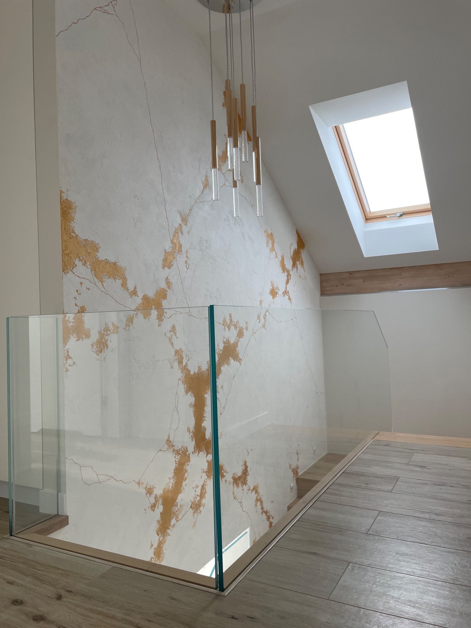 Ściana wewnątrz domu z dekoracyjnym tynkiem imitującym spękania złota, oświetlona wiszącą lampą i światłem z okna dachowego, zabezpieczona szklaną balustradą.