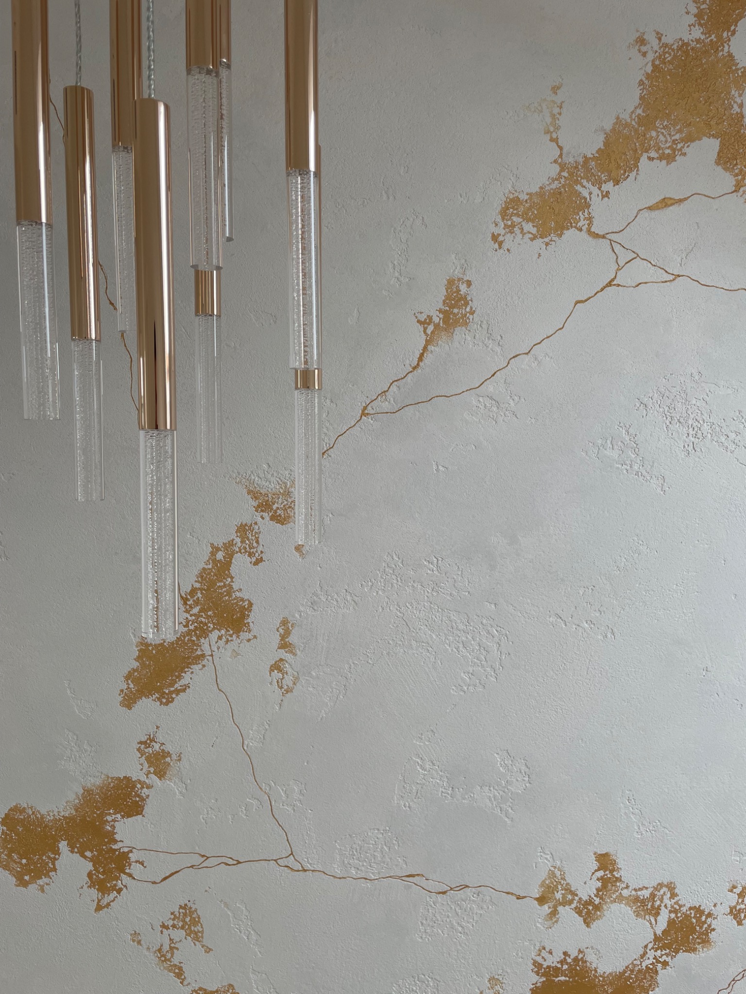 Dekoracyjna ściana z efektem spękań i złotymi akcentami, uzupełniona nowoczesnym oświetleniem w postaci długich, przezroczystych tub z elementami w kolorze złota.