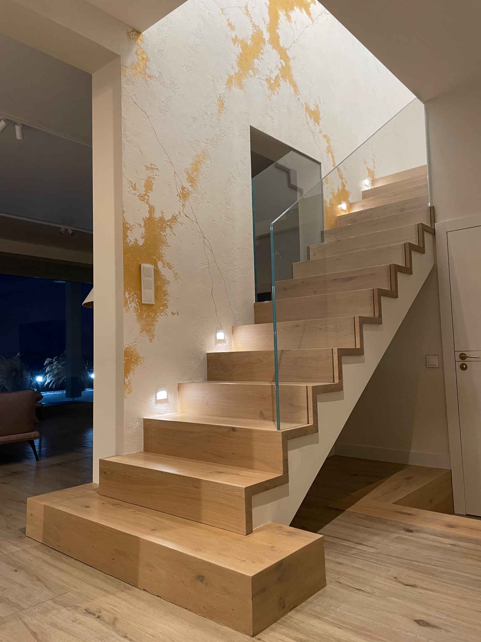 Nowoczesne drewniane schody z szklaną balustradą i oświetleniem LED w ścianie, na ścianie dekoracyjna faktura z elementami złotej farby.