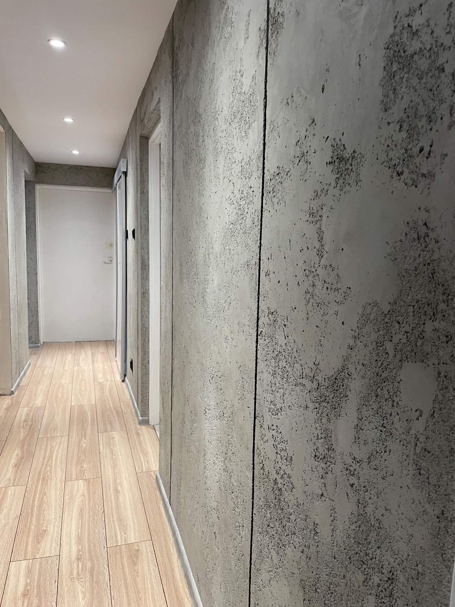 Korytarz z betonową ścianą o nieregularnej fakturze, imitującej surowy beton, jasna podłoga z paneli imitujących drewno, białe drzwi w głębi kadru.