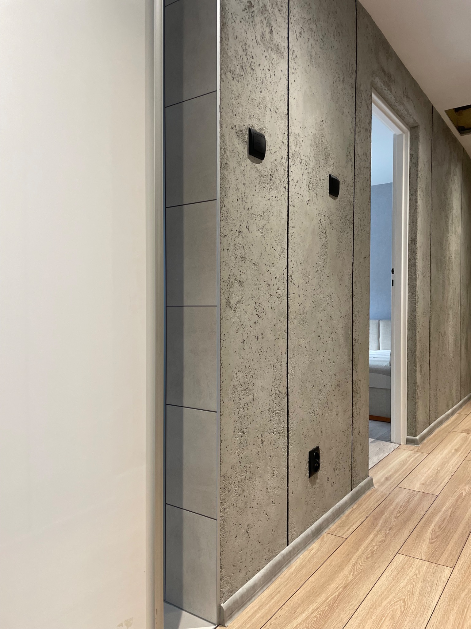 Korytarz z wykończeniem ścian w stylu surowego betonu, z widocznymi pionowymi podziałami, czarnymi gniazdkami elektrycznymi i panelami podłogowymi w odcieniach drewna. Po lewej stronie białe drzwi,...