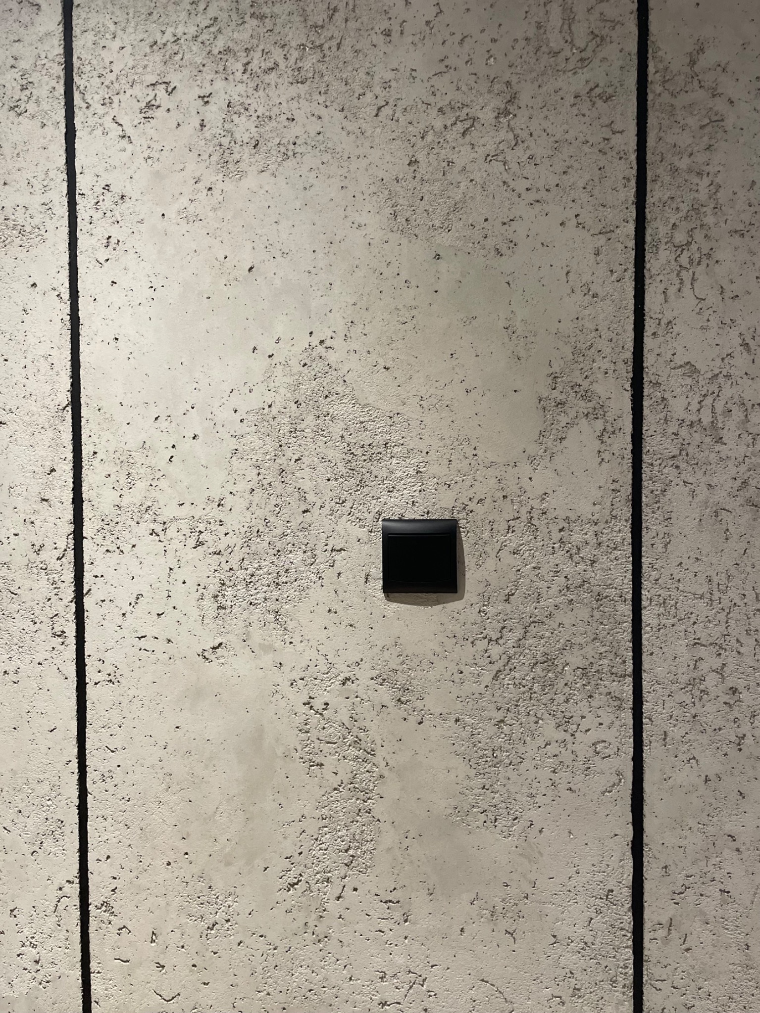 Szara ściana z wyraźną fakturą i pionowymi, ciemnymi liniami, z zamontowanym czarnym włącznikiem światła po lewej stronie.