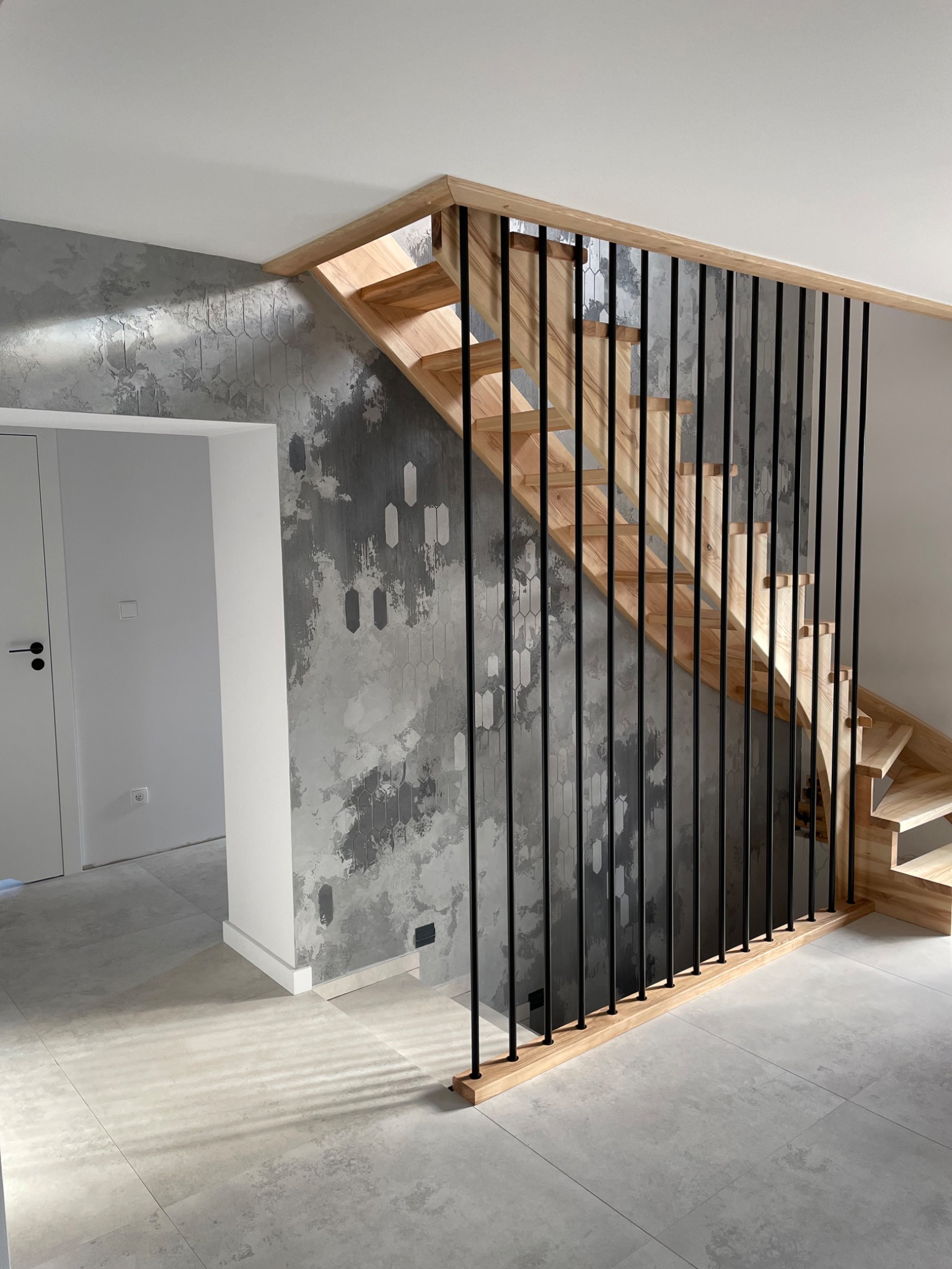 Nowoczesne schody drewniane z czarnymi, pionowymi balustradami na tle ściany z dekoracyjnym tynkiem imitującym beton z geometrycznym wzorem, widok na białe drzwi i jasną podłogę z płytek.