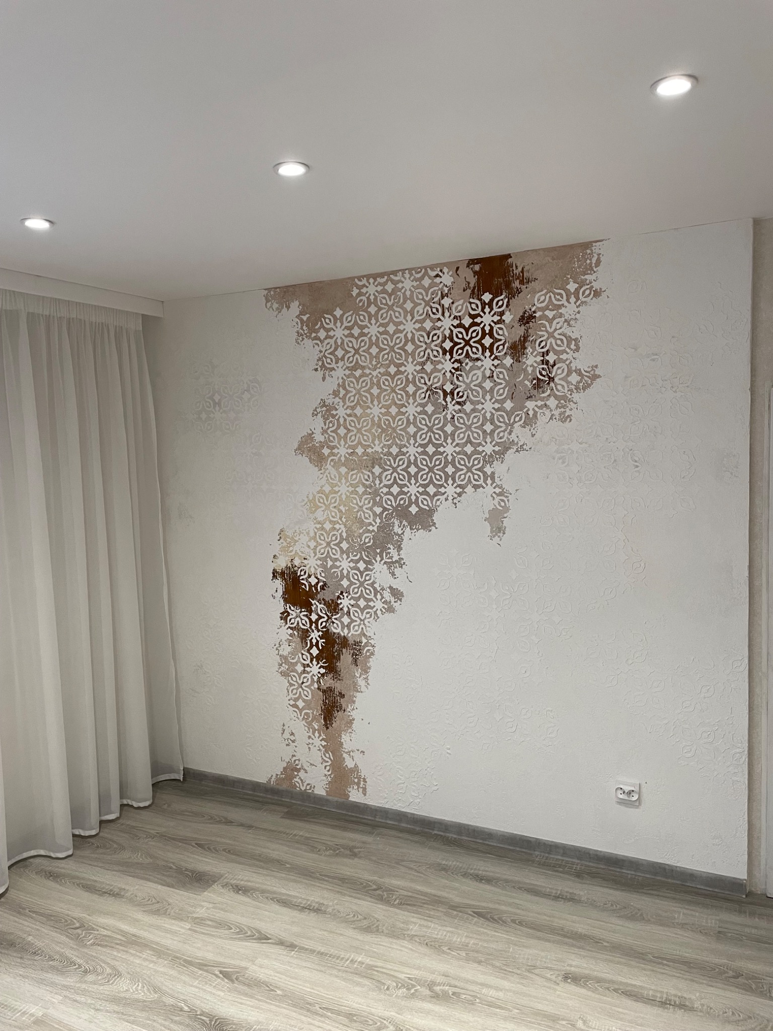 Ściana w pokoju z dekoracyjnym wzorem kwiatowym w odcieniach brązu i bieli, częściowo pokrywającym teksturowaną powierzchnię. Widoczne szare panele podłogowe i białe zasłony.